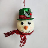 A handcrafted snowman ornament with a green hat and red scarf, hanging from a string.